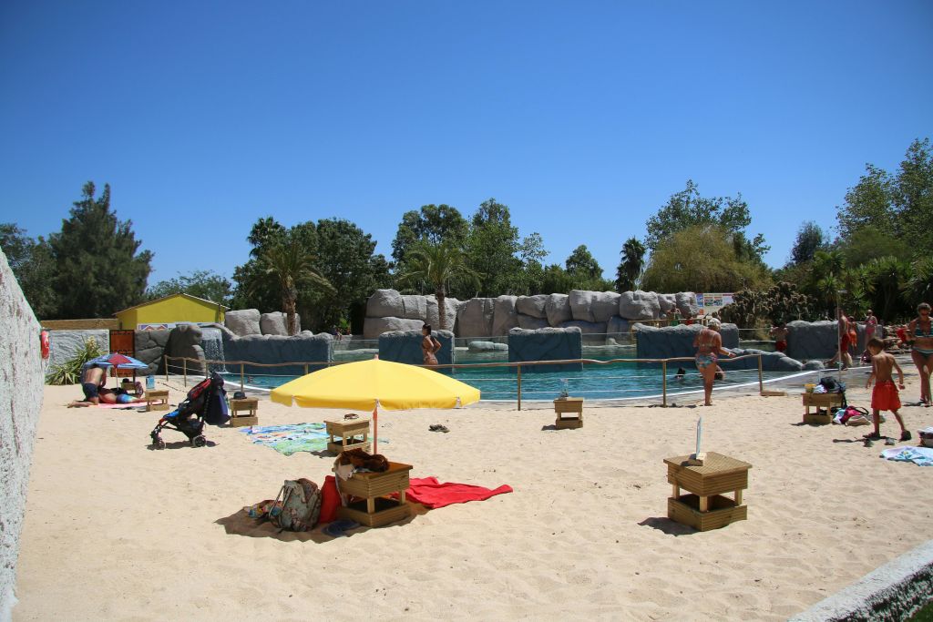 라운지 공간과 커다란 수영장이 내려다보이는 노란 우산이 있는 모래사장. 사람들이 걷고 있고 유모차와 가방이 있습니다.