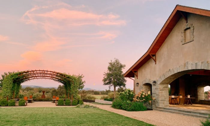 커다란 집, 정원 아치길, 벤치, 나무, 파스텔빛 노을 하늘 아래 광활한 잔디밭이 있는 매력적인 시골 풍경.