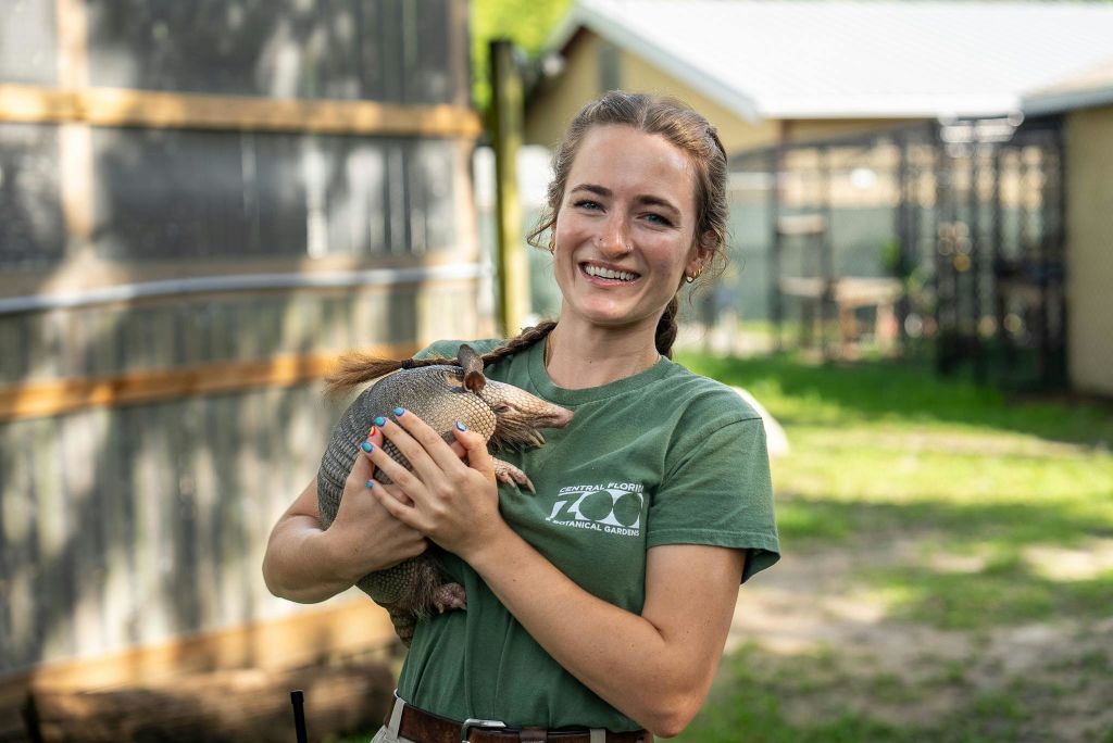 야외에서 미소를 짓고 아르마딜로를 들고 동물원 로고가 있는 녹색 티셔츠를 입은 사람. 배경은 건물과 녹지를 보여줍니다.
