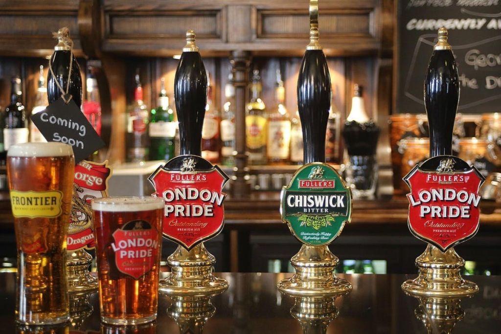 풀러스 런던 프라이드 (Fuller's London Pride) 와 치스윅 비터 (Chiswick Bitter) 라는 이름의 맥주 탭이 있는 펍 카운터와 카운터에 맥주 한 잔이 있습니다.