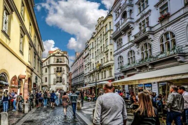 부분적으로 흐린 하늘 아래 화려한 건물과 야외 카페가 늘어선 유럽 거리를 걷는 보행자.