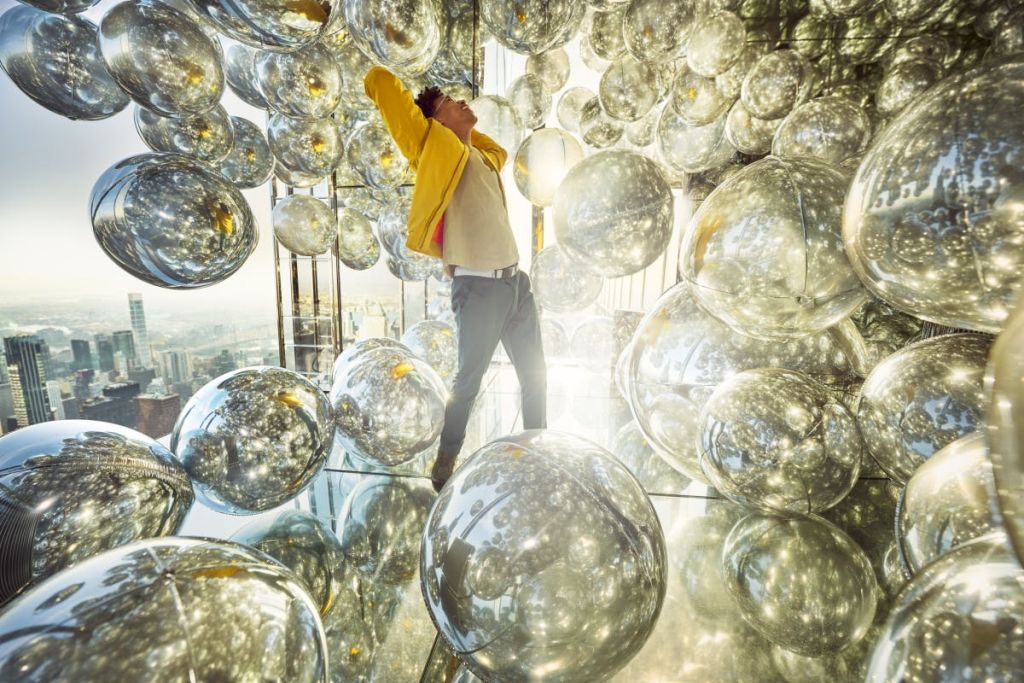 한 사람이 도시 바깥 풍경을 볼 수 있는 조명이 밝은 방에서 팔을 들고 뻗어 수많은 반사 은색 구체들 사이에 서 있습니다.