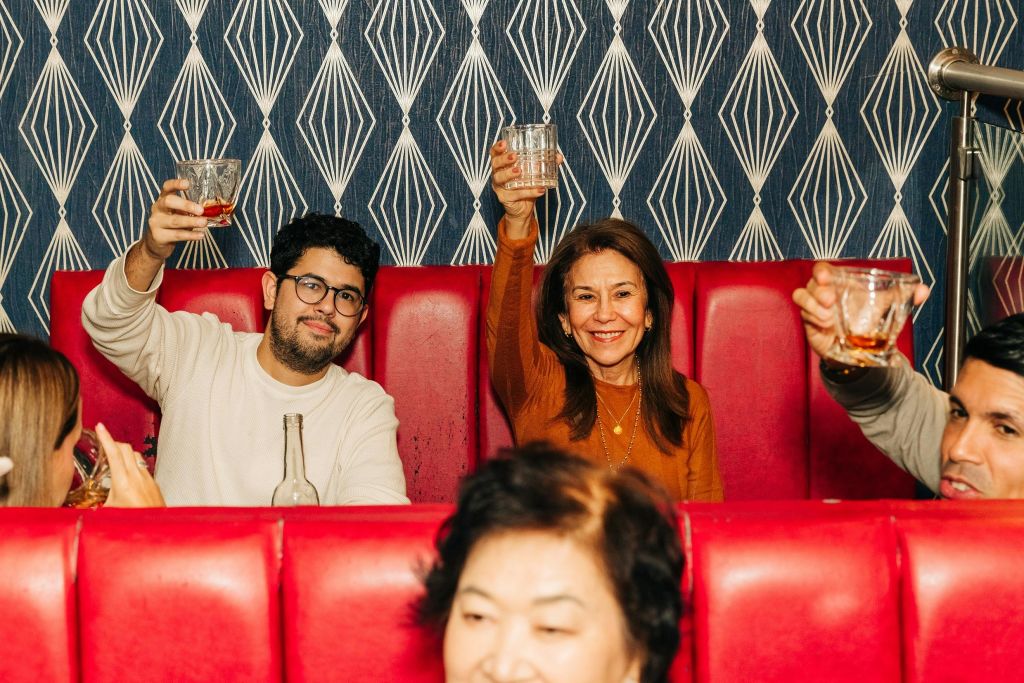 빨간색 좌석이 있는 부스에 세 사람이 앉아 미소를 지으며 안경을 들어 올리고 있습니다. 배경에 무늬가 있는 벽지가 보입니다.