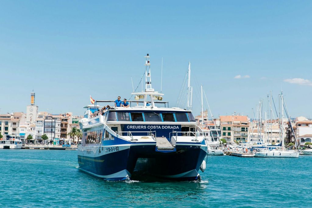 건물과 요트를 배경으로 항구 근처의 푸른 바다를 항해하는 '크루어스 코스타 도라다 (Creuers Costa Dorada)' 라는 이름의 페리.