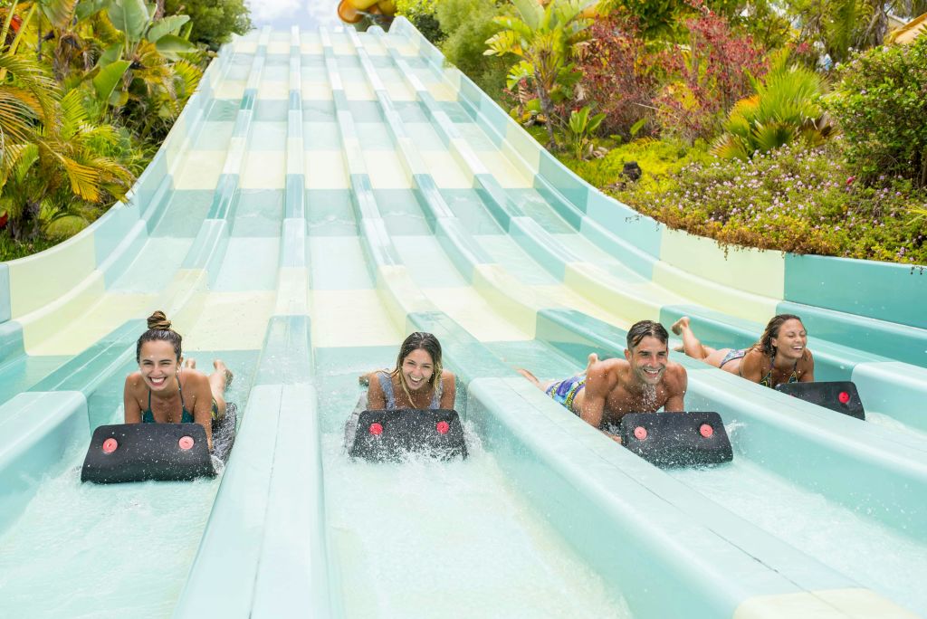 세 사람이 매트 위에서 평행한 워터 슬라이드를 미끄러지며 미소를 짓고 울창한 녹음으로 둘러싸여 있습니다.