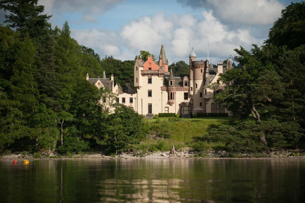 푸르른 녹음으로 둘러싸인 포탑과 가파른 지붕이 있는 크림색의 성은 잔잔한 물에 반사됩니다.