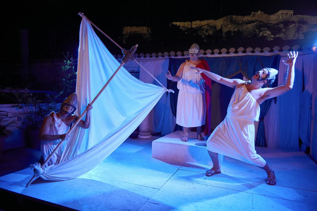 고대 그리스 의상을 입은 세 명의 배우가 파란색 조명 아래에서 극적인 포즈와 표정으로 야외 무대에서 공연합니다.