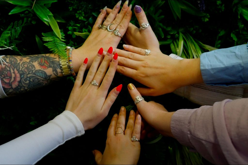 다양한 반지와 네일 컬러를 가진 다섯 개의 손이 녹색 잎을 배경으로 원을 그리며 뻗어 있습니다.