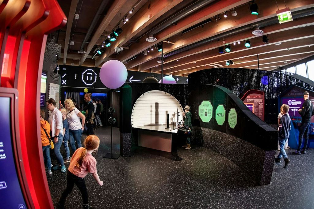 양방향 과학을 탐구하는 사람들은 조명, 그림자, 디스플레이 보드로 전시합니다. 분홍색 셔츠를 입은 소녀가 전경을 걷고 있습니다.