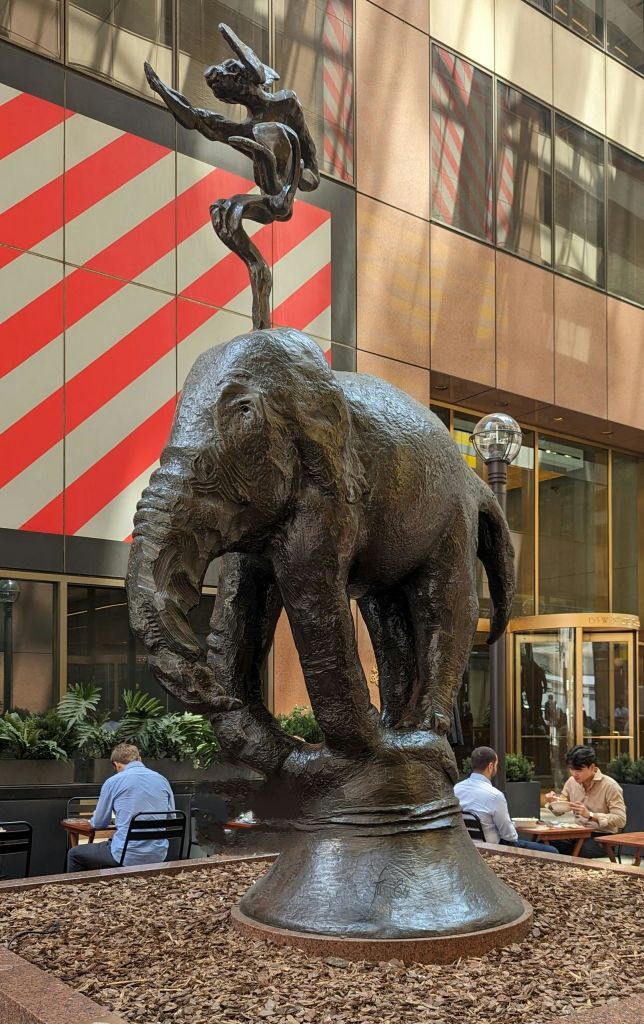 야외에는 청동 코끼리 조각상이 전시되어 있으며, 근처에 사람들이 앉아 식사를 하고 있습니다. 건물이 배경에 있습니다.