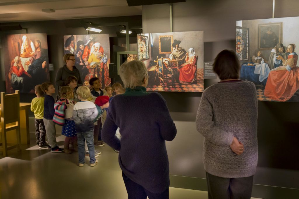 어린이와 성인은 박물관에서 그림을 관찰하며, 벽에 역사적인 장면을 묘사한 세부 예술 작품을 감상할 수 있습니다.