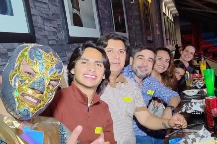 조명이 어두운 레스토랑의 테이블에 여섯 명이 앉아 미소를 지으며 카메라를 향해 포즈를 취하고 있습니다.