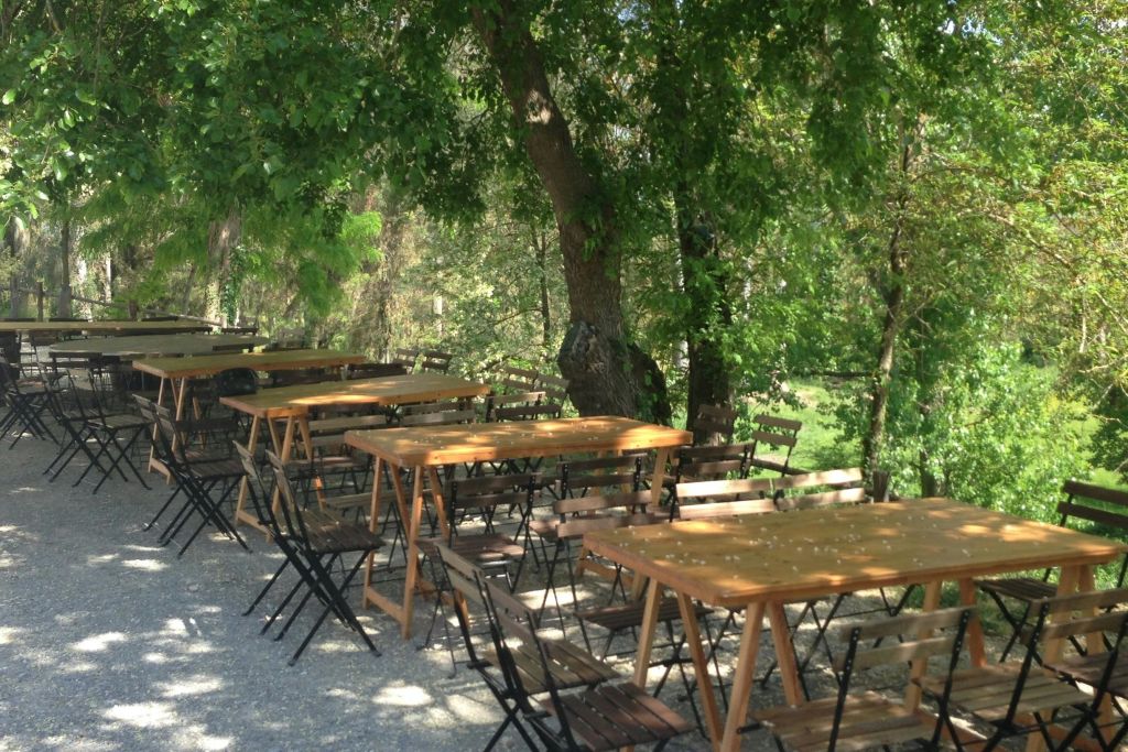 잎이 무성한 커다란 나무 아래 자갈 표면에 나무 테이블과 의자가 야외에 배치되어 있습니다.