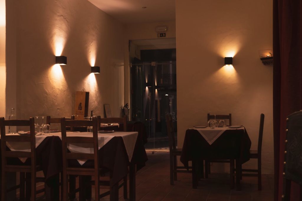 나무 테이블과 의자, 흰색 식탁보, 벽걸이 조명으로 따뜻한 분위기를 연출하는 조명이 희미한 레스토랑입니다.