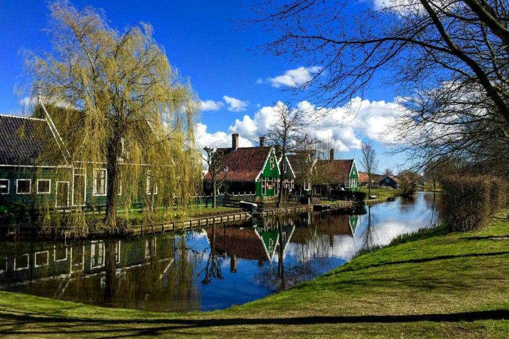 흩어져 있는 구름과 함께 맑고 푸른 하늘 아래, 나무 옆으로 펼쳐진 고요한 운하에 전통 녹색 목조 주택이 반사됩니다.