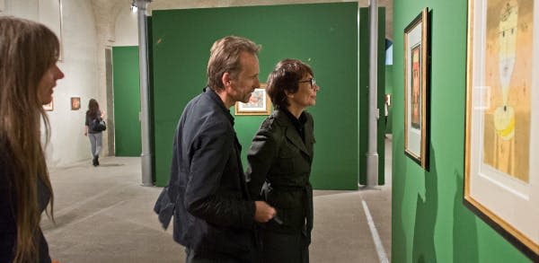 어두운 옷을 입은 두 사람이 실내 갤러리의 녹색 벽에 걸린 액자 예술을 관찰합니다.