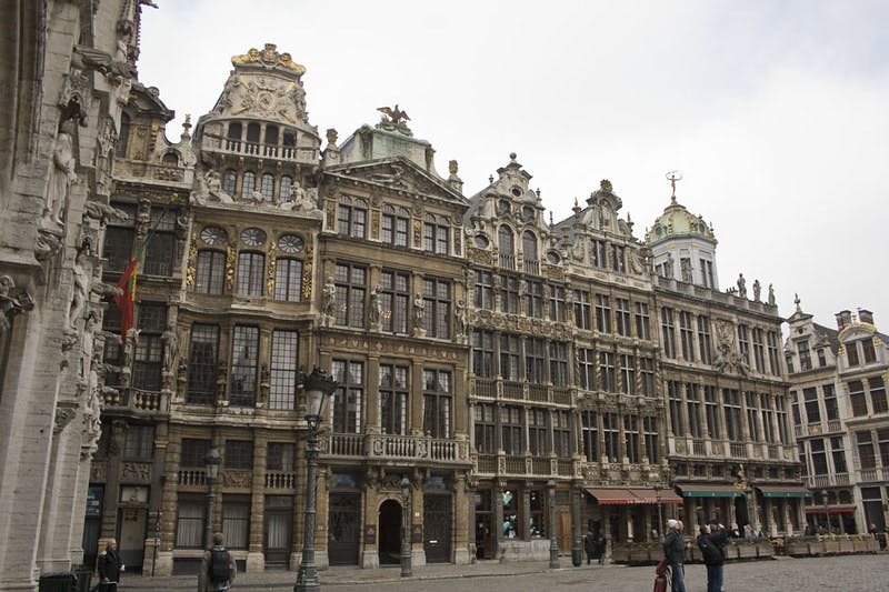 자갈길에 디테일한 외관이 있는 화려한 역사적 건물이 있습니다. 보행자 몇 명이 걸어가고 있습니다.