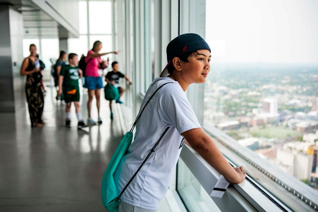 흰색 셔츠와 야구 모자를 쓴 소년이 도시 풍경을 배경으로 고층 건물의 커다란 유리창을 바라보고 있습니다.