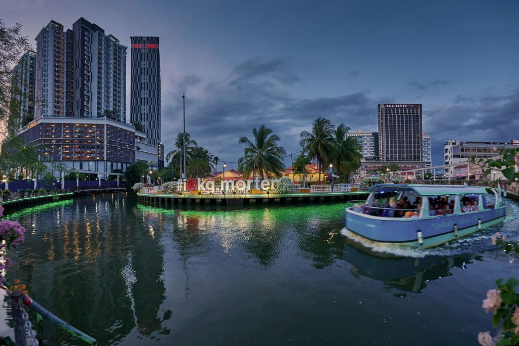 야자수, 건물, 네온 조명 "Kampung Morten" 표지판이 있는 해질 무렵의 수변 지역. 전경의 강가에 있는 보트.