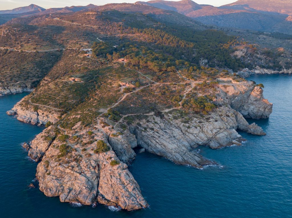 험준한 지형, 흩어져 있는 초목, 바다로 이어지는 구불구불한 길이 있는 바위 해안의 공중 전망. 산을 배경으로.