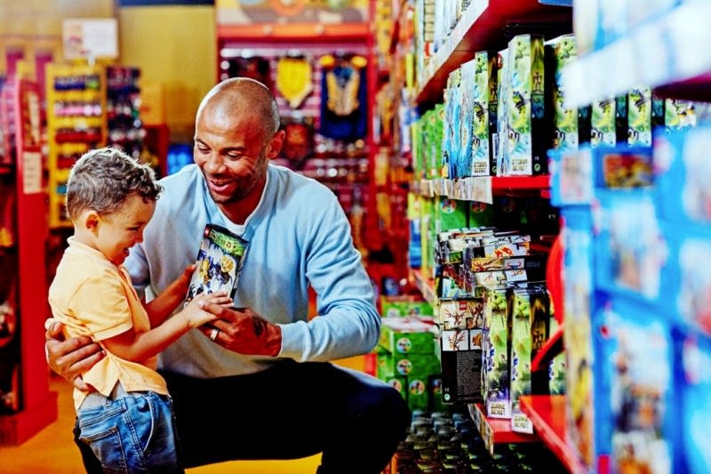한 남자와 어린이가 다채로운 장난감 가게 통로에서 즐겁게 장난감 패키지를 보고 있습니다.