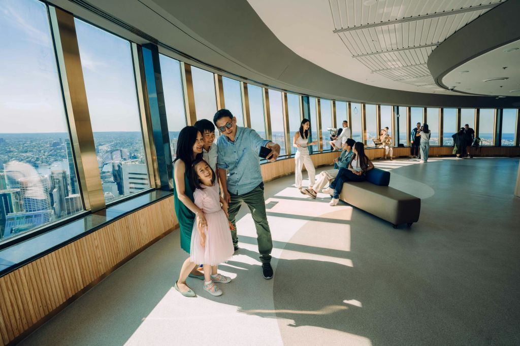 4인 가족이 커다란 창문이 있는 현대적인 전망대에서 셀카를 찍으며 도시의 스카이라인을 감상할 수 있습니다.