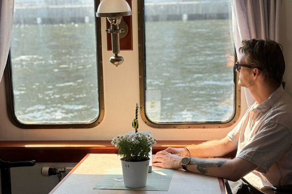 문신이 새겨진 팔을 가진 사람이 화분이 있는 테이블에 앉아 있습니다. 창문 옆에는 물이 내려다보입니다.