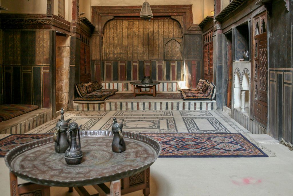 쿠션이 있는 휴식 공간, 중앙의 낮은 테이블, 둥근 트레이 위에 놓인 금속 주전자, 무늬가 있는 러그가 있는 화려하게 꾸며진 객실입니다.