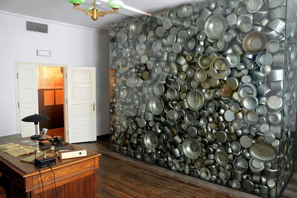 문이 열린 방, 나무 책상, 녹색 천장 조명이 있는 방에는 금속 냄비와 프라이팬으로 덮인 벽이 있습니다.