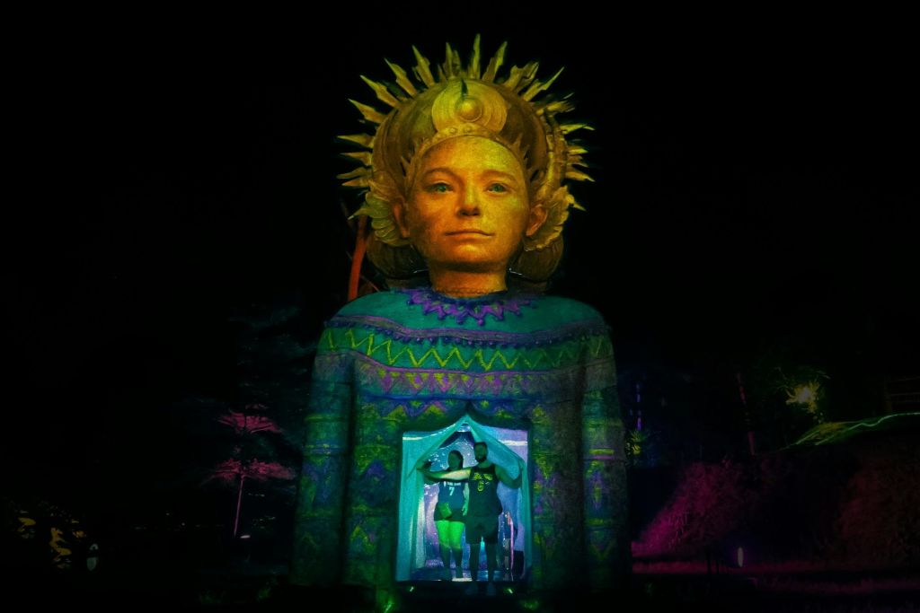 밤에 황금빛 햇살이 내리쬐는 머리 장식이 있는 커다란 조명 동상. 두 사람이 다채로운 무늬로 둘러싸인 기슭의 아치형 통로에 서 있습니다.