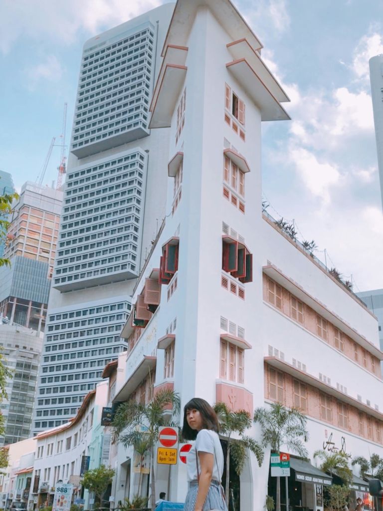 부분적으로 흐린 하늘 아래 높고 현대적이며 오래된 건물이 있는 길 모퉁이에 한 여성이 서 있습니다.