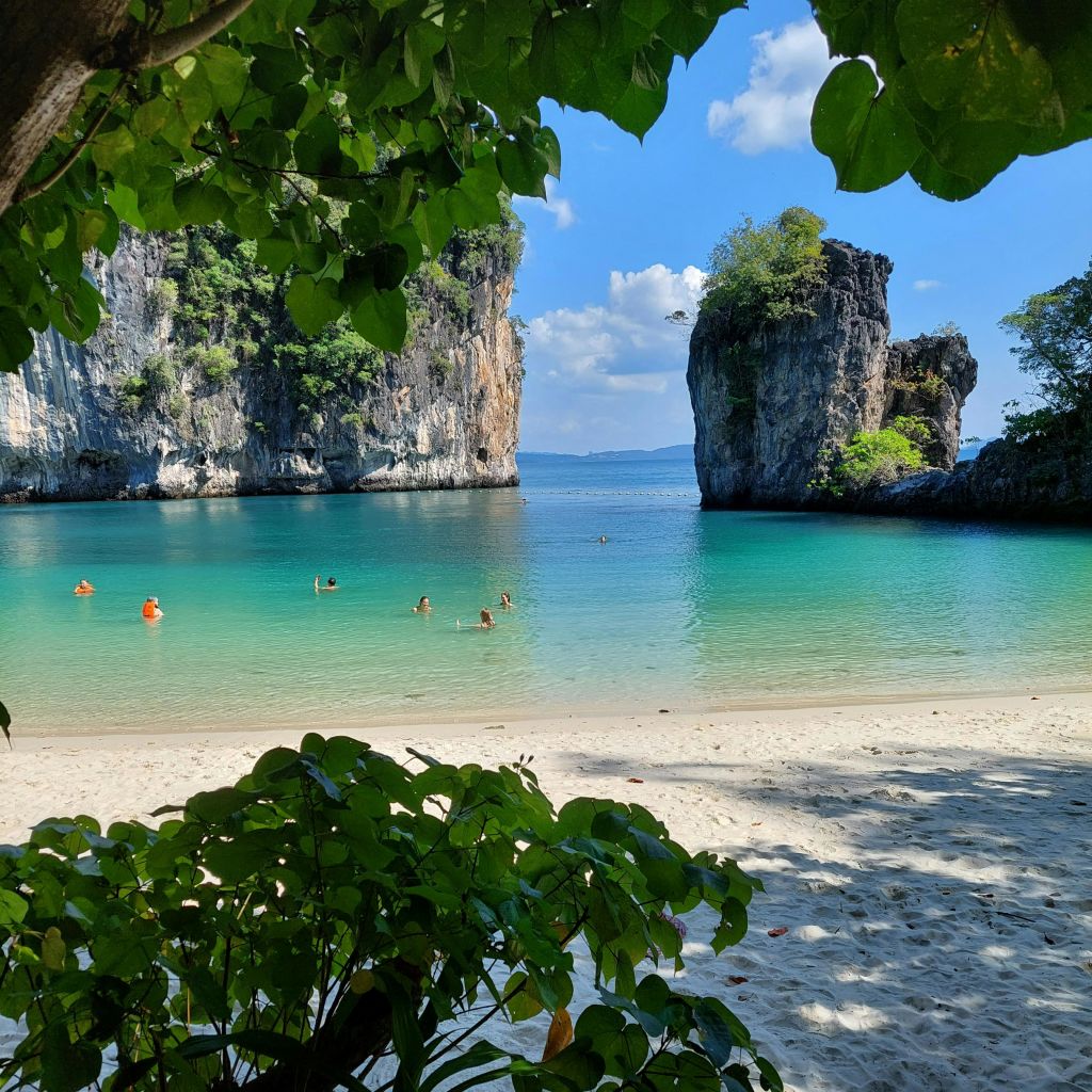 바위 절벽과 녹음으로 둘러싸인 청록색 물에서 수영하는 사람들로 모래 해변과 잎이 우거진 나무 캐노피가 눈앞에 펼쳐집니다.