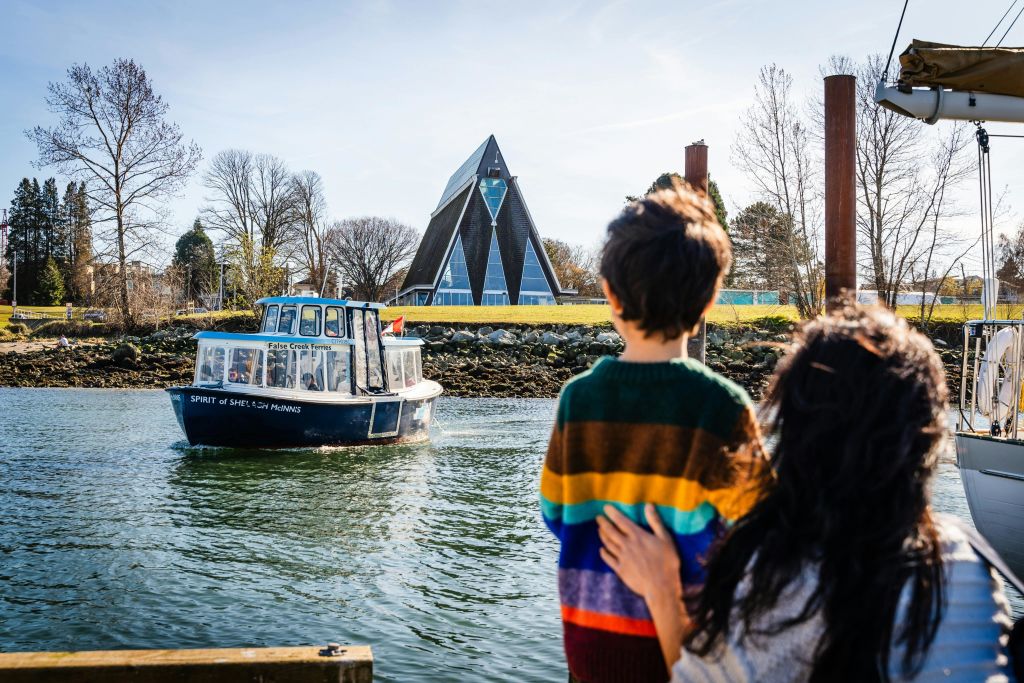 한 사람과 어린이가 작은 배를 보며 물가에 서 있습니다. 배경에는 현대적인 삼각형 건물이 보입니다.