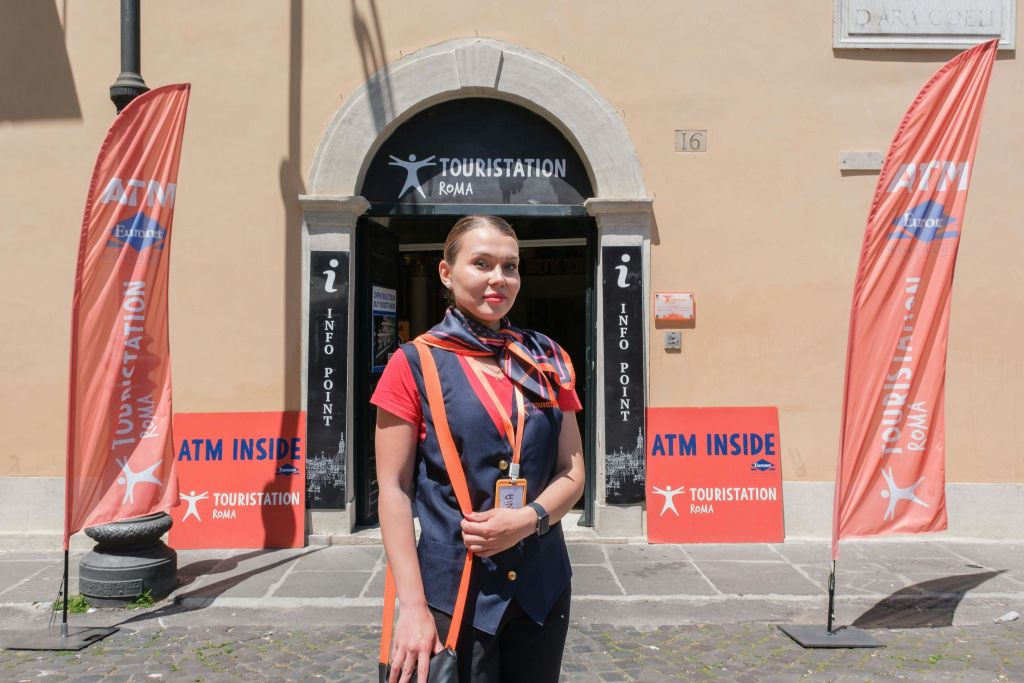 "Touristation Roma" 라고 표시된 건물 앞에 유니폼을 입은 여성이 서 있으며 안내 표지판과 내부의 ATM을 알리는 표지판이 있습니다.