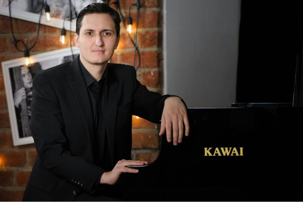 벽돌 벽에 걸린 조명과 액자 사진을 배경으로 피아노 옆에서 포즈를 취하는 정장 차림의 사람.