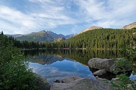 나무가 늘어선 해안과 부분적으로 흐린 하늘 아래 험준한 봉우리가 선명하게 반사된 고요한 산악 호수.