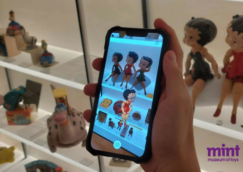 실제 베티 부프 인형 두 개를 배경으로 베티 부프 인형의 증강 현실 이미지가 표시된 스마트폰을 들고 있는 손.