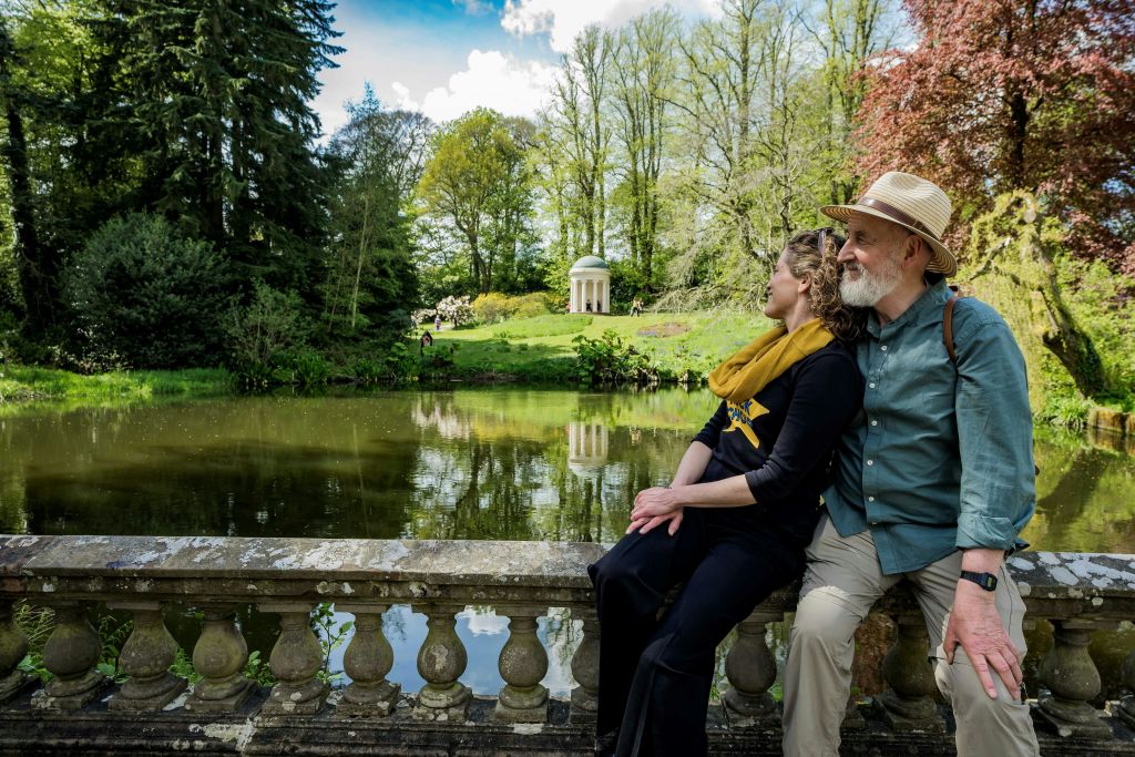 울창한 정원의 연못 옆 석조 난간에 노부부가 앉아 있습니다. 배경에는 작은 사원이 보입니다.