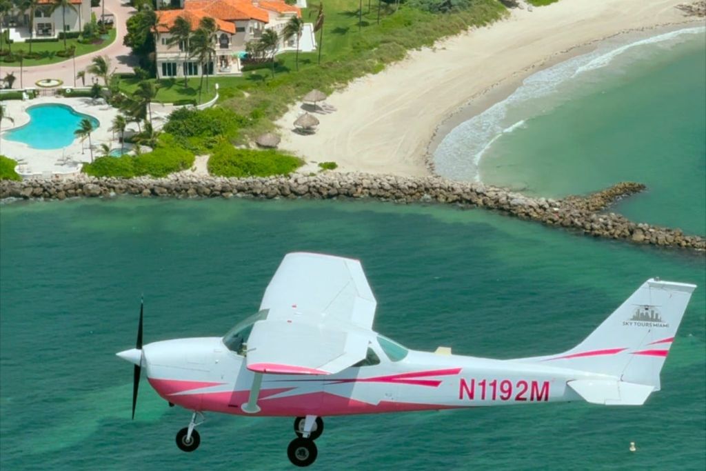 하얀색과 분홍색 프로펠러 비행기가 집과 야자수를 배경으로 모래 해변 근처의 청록색 물 위로 날아갑니다.