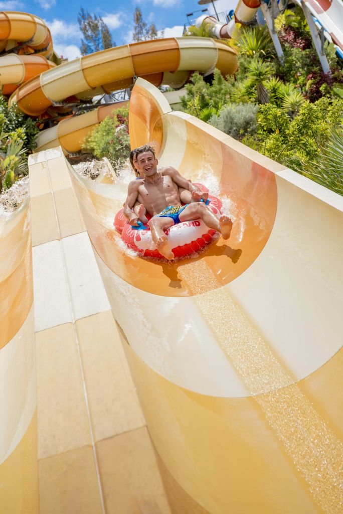 무성한 녹음과 복잡한 슬라이드 구조를 배경으로 물 미끄럼틀 아래로 팽창식 뗏목을 타는 사람.