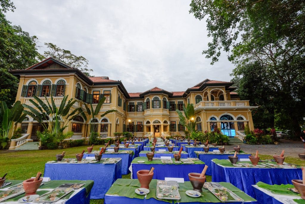 커다란 노란색 식민지 풍의 건물 앞에 초록색 덮개와 진흙 모르타르, 해충이 놓인 테이블이 줄지어 있습니다.