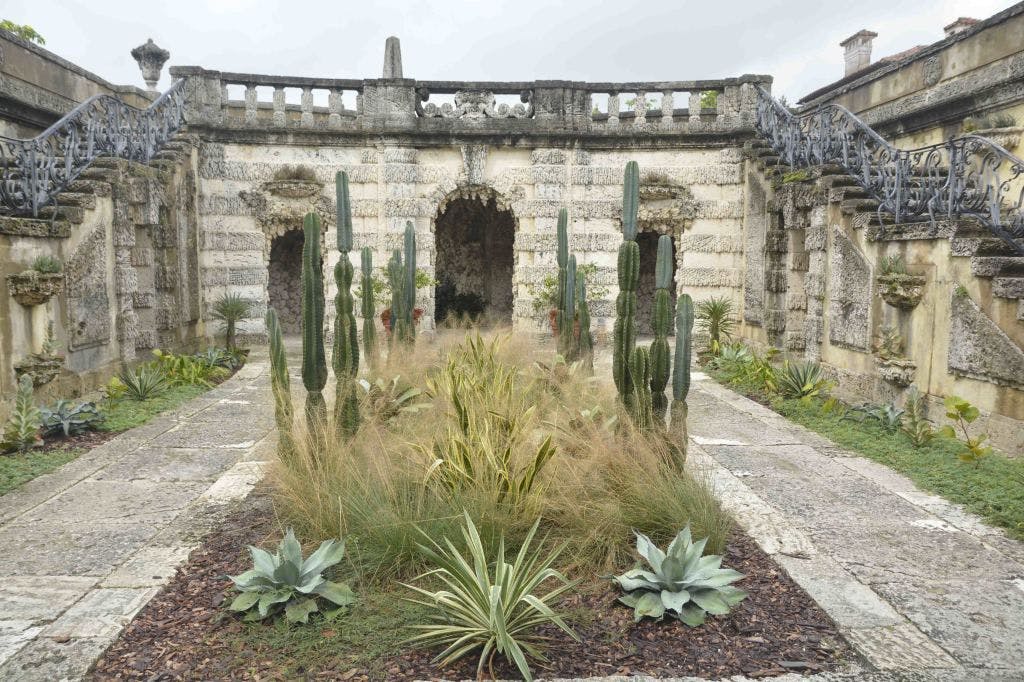 높은 선인장, 장식용 잔디, 아치형 출입구와 난간이 있는 풍화된 석조 건물이 있는 석조 안뜰.