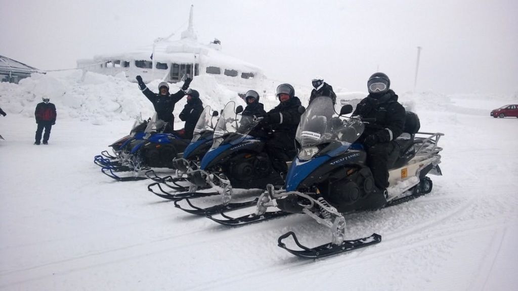 눈 덮인 건물을 배경으로 눈 덮인 풍경의 파란색과 흰색 스노우모빌에 앉아 있는 5명의 겨울맞이 장비를 착용한 사람들.