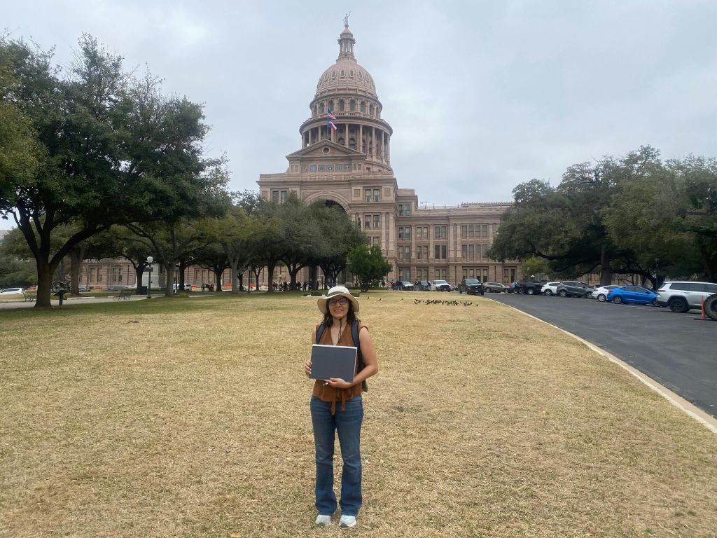 한 사람이 노트북을 들고 잔디밭에 서 있습니다. 커다란 돔형 건물이 배경으로 흐린 하늘 아래 있습니다.