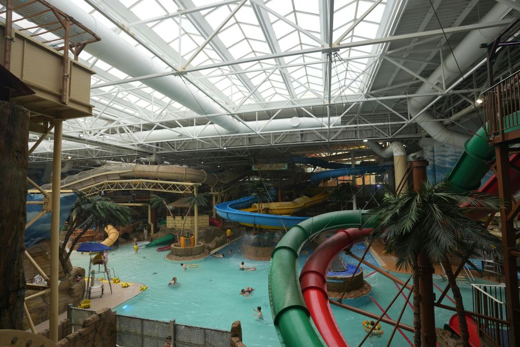 수영장, 워터슬라이드, 야자수, 금속 기둥이 있는 높은 천장 아래에서 수영하는 사람들이 있는 실내 워터파크.