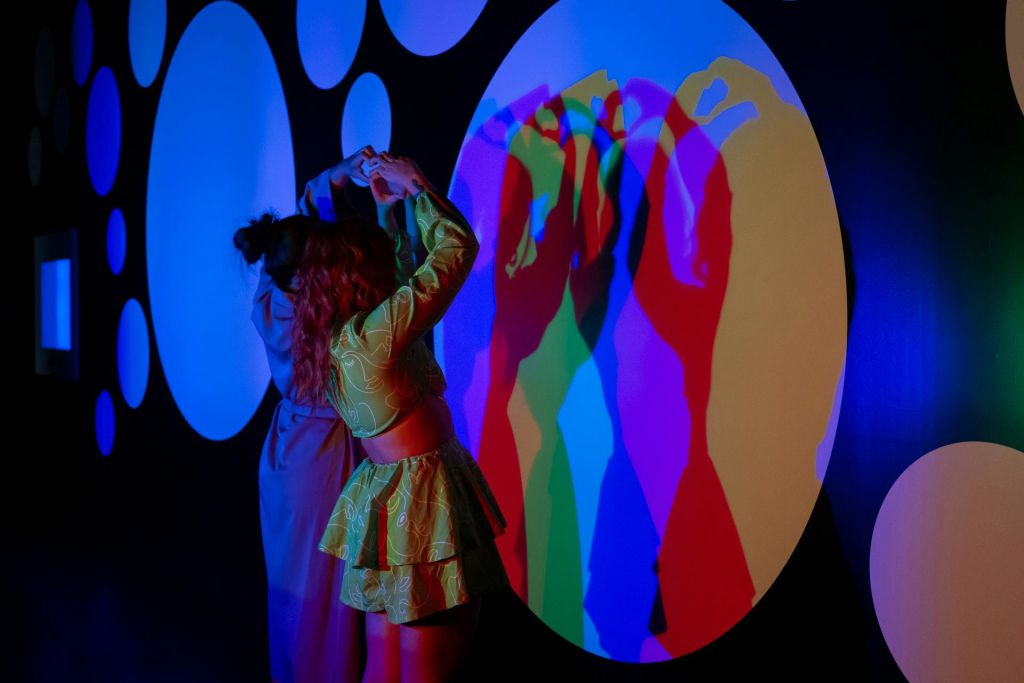 두 사람은 파란색, 녹색, 빨간색, 보라색 조명이 겹쳐진 다채로운 색상의 벽면 디스플레이에서 그림자 모양을 만듭니다.