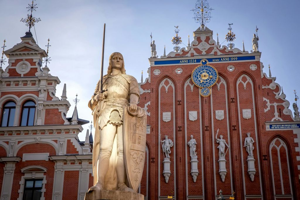 섬세한 디테일과 시계가 돋보이는 화려하고 유서 깊은 건물 앞에 검과 방패를 든 기사의 동상이 서 있습니다.