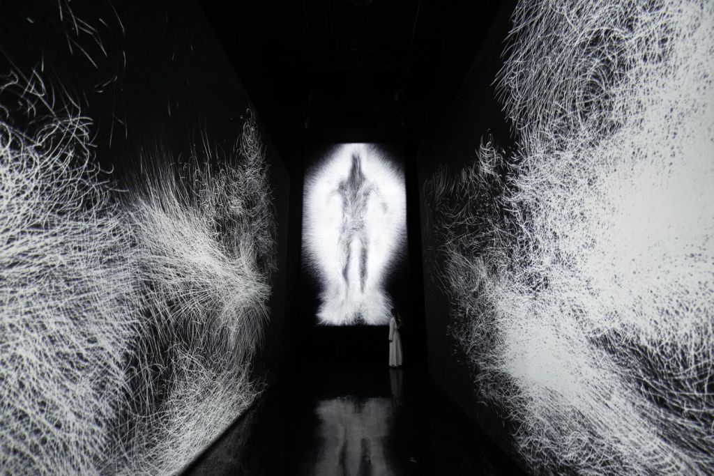 벽에 추상적인 흰색 무늬가 있는 어두운 복도와 그 끝에 빛나는 사람 같은 형상이 투영되어 있습니다.