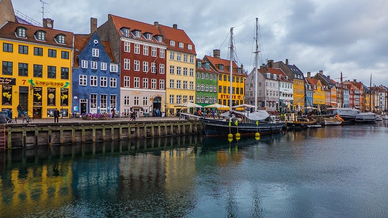 흐린 하늘 아래 돛단배가 정박해 있는 바닷가를 따라 화려한 건물들이 줄지어 있습니다.