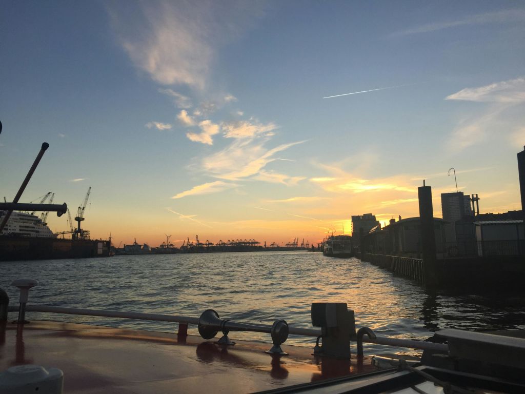 부분적으로 흐린 하늘을 배경으로 실루엣으로 크레인, 보트, 산업용 건물이 있는 항구 위로 석양을 감상해보세요.